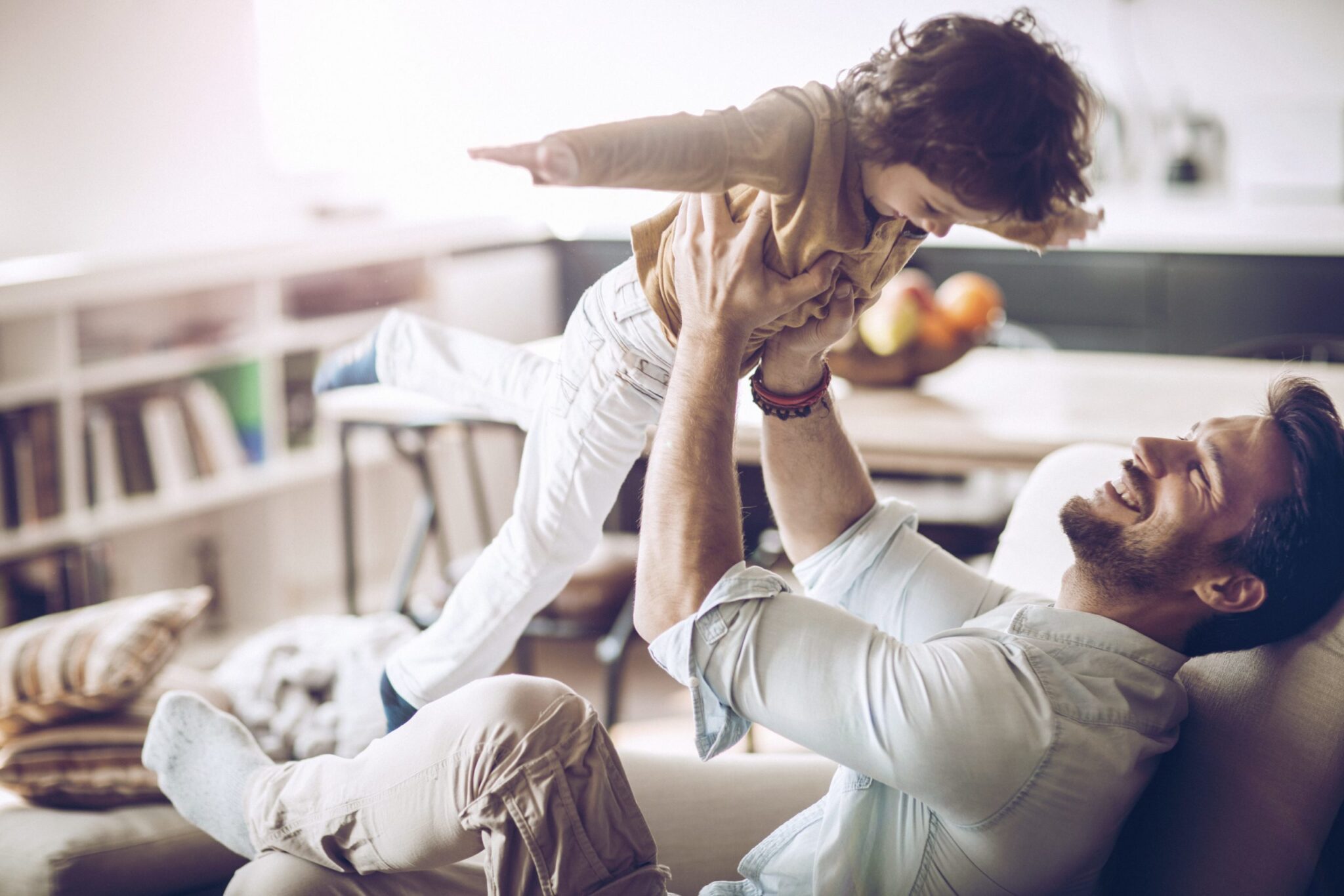 child with curly brown hair being held up in the air by his dad laughing